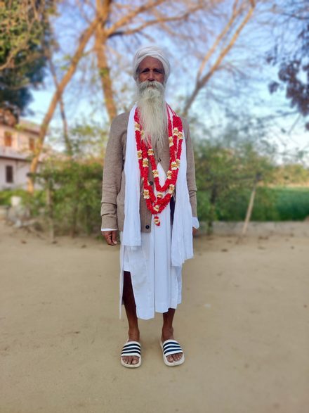Sant Baba Jagjit Singh Ji standing outdoors in white, with a white turban, a fawn cardigan and a red garland